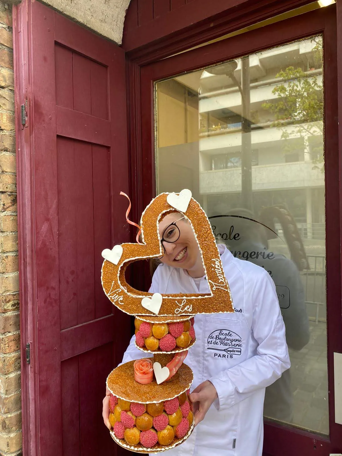 Laura Bilbault en veste de pâtissière devant l'École de Boulangerie et de Pâtisserie de Paris, portant un croquembouche en forme de cœur